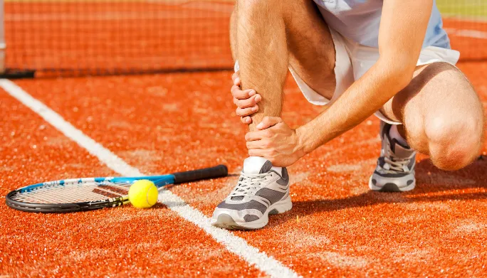 Injured tennis player on red clay court, holding ankle near tennis racket and ball, highlighting sports rehabilitation needs.